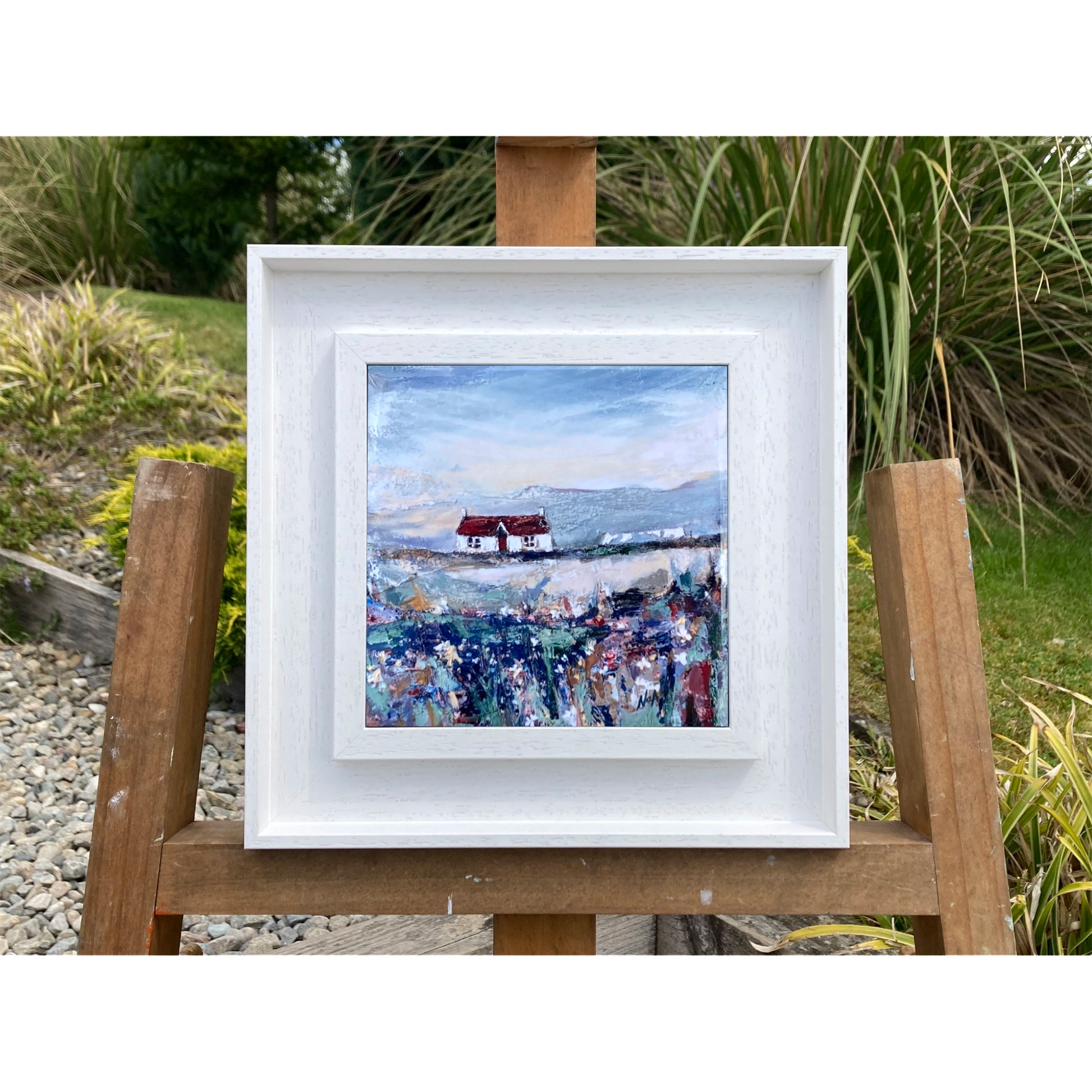 A ceramic tile printed with an abstract painting of a cottage and flowers mounted on a white frame