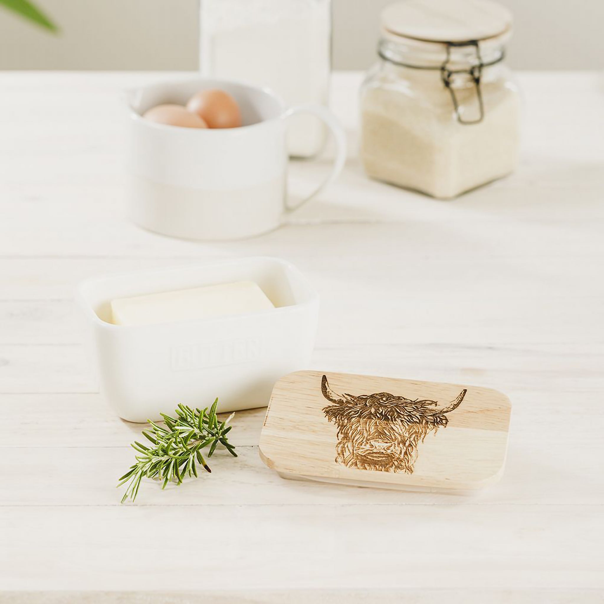 White ceramic butter dish with a wooden lid engraved with a Highland cow staged on a table with butter and rosemary