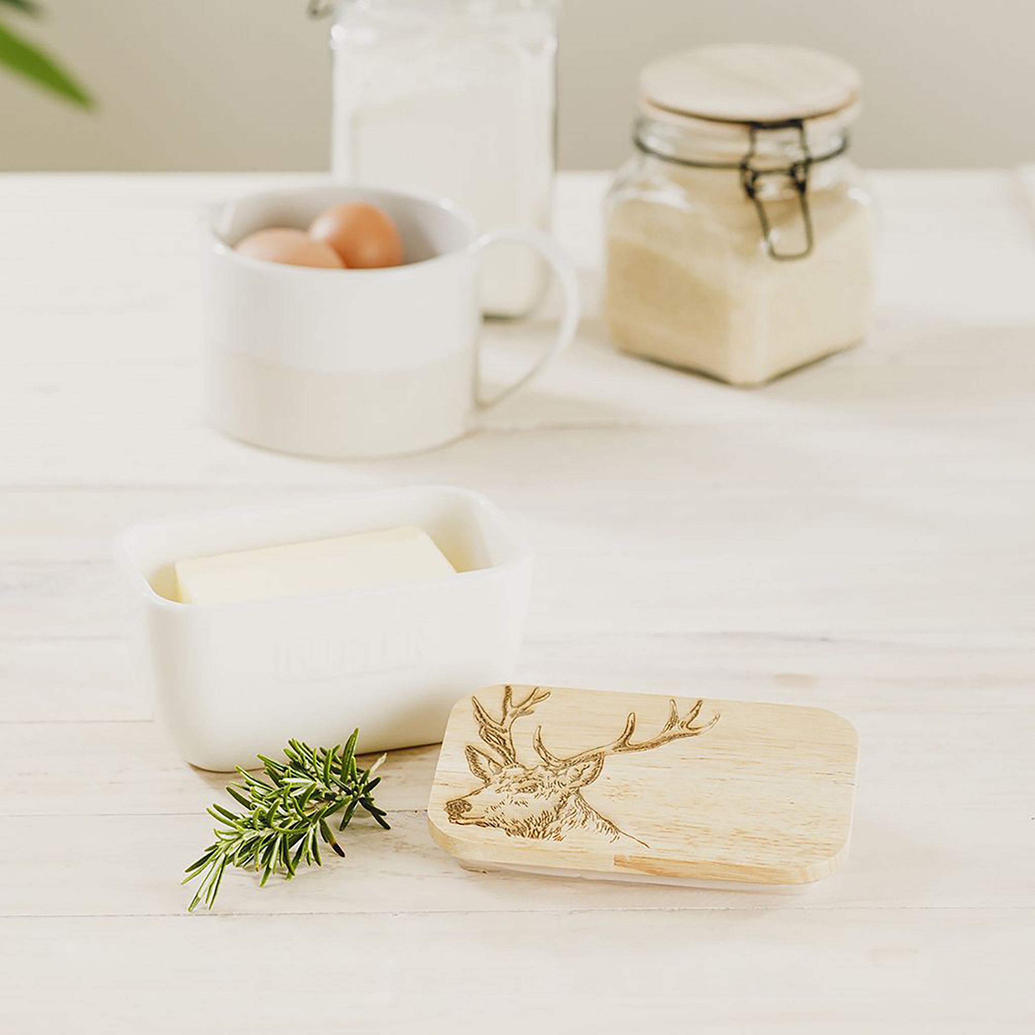 White ceramic butter dish with a wooden lid engraved with a stag staged on a table with butter and rosemary