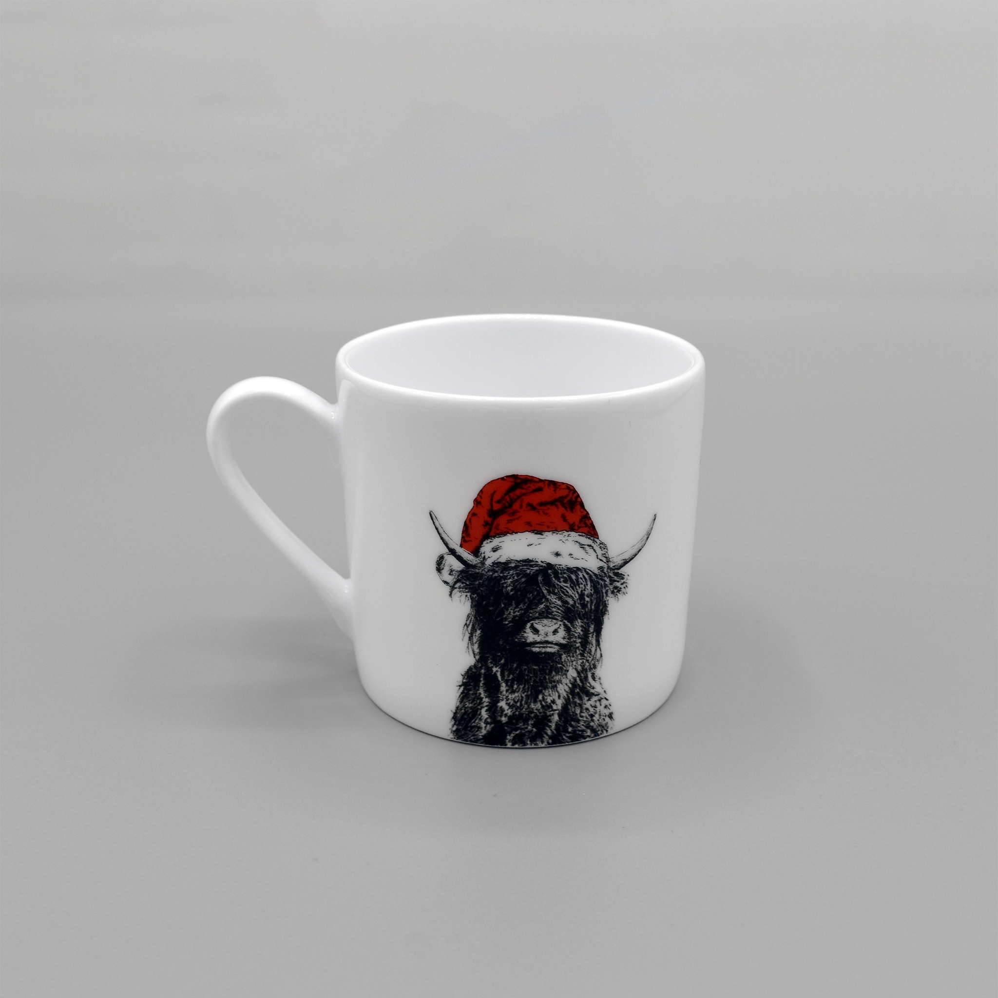 A white espresso mug featuring a printed image of a Highland cow wearing a red Santa hat