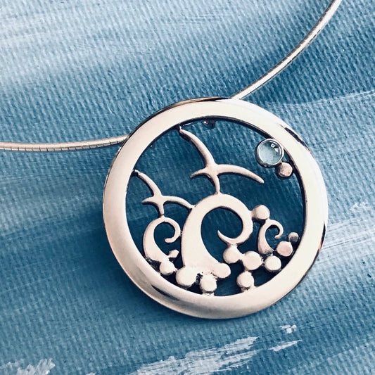 A large round silver pendant on a neck wire featuring a frame with flying birds, tall curled waves and a blue topaz