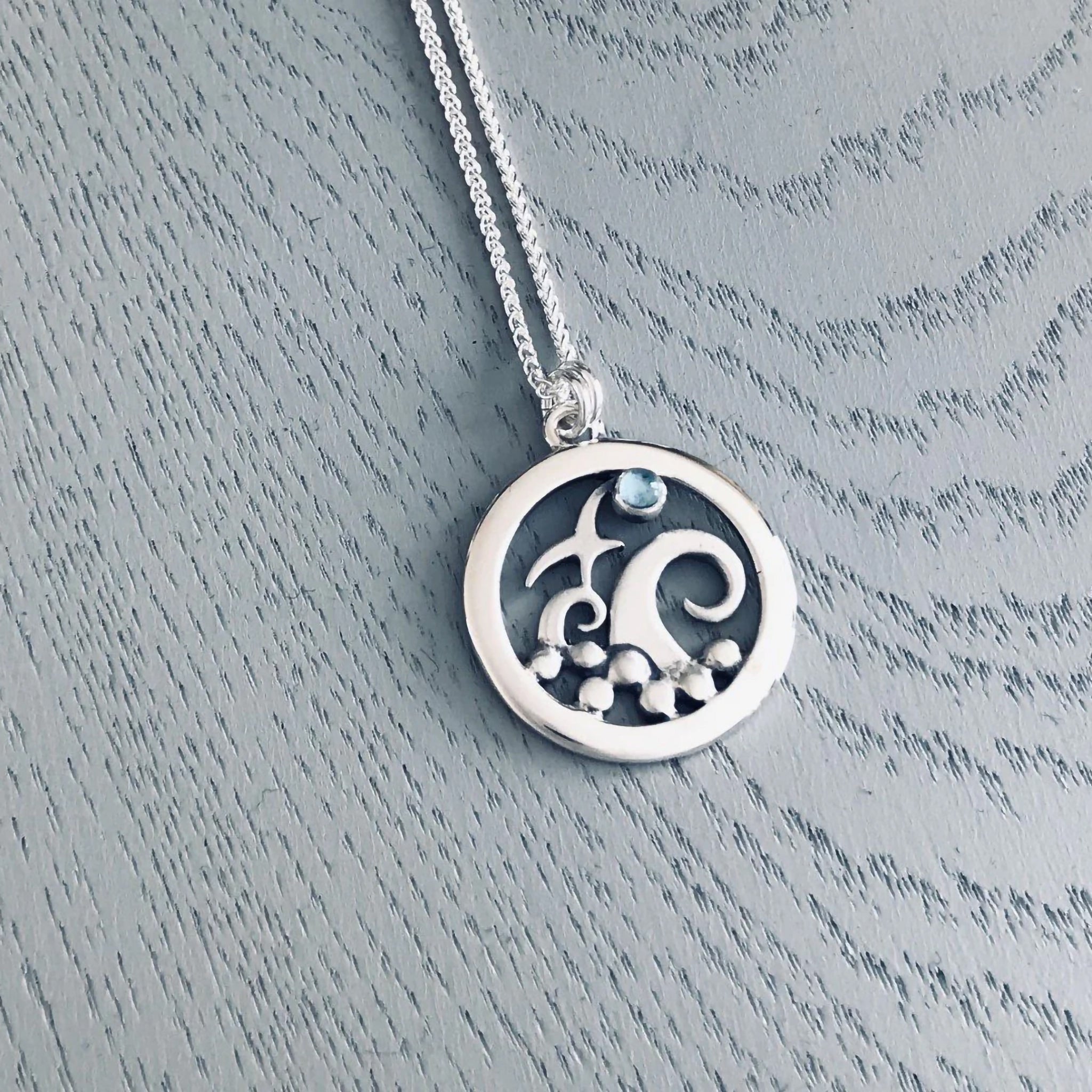 A round silver pendant featuring a bird with tall curled waves and a round blue topaz stone