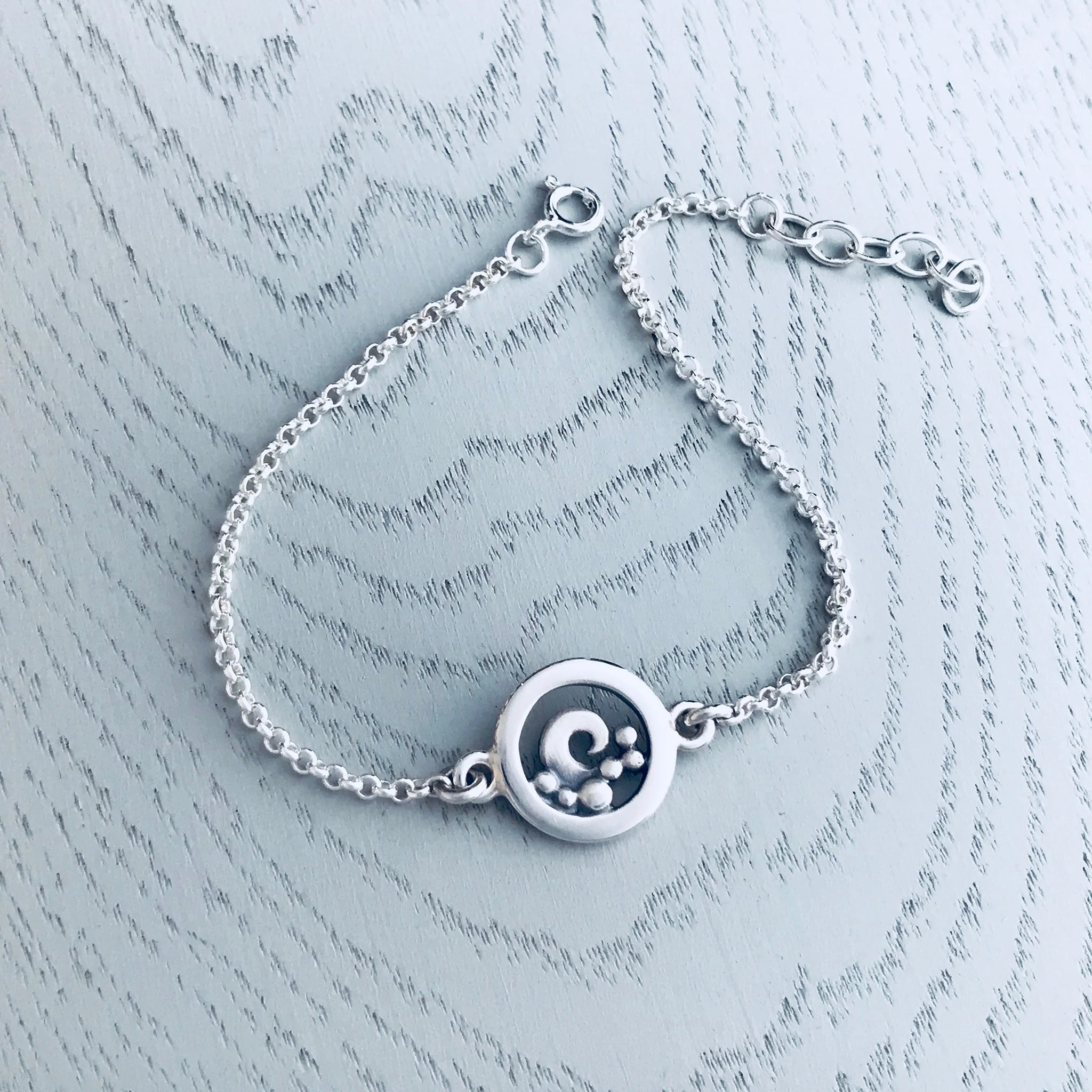 A silver chain bracelet featuring a round pendant with a frame and curled wave design