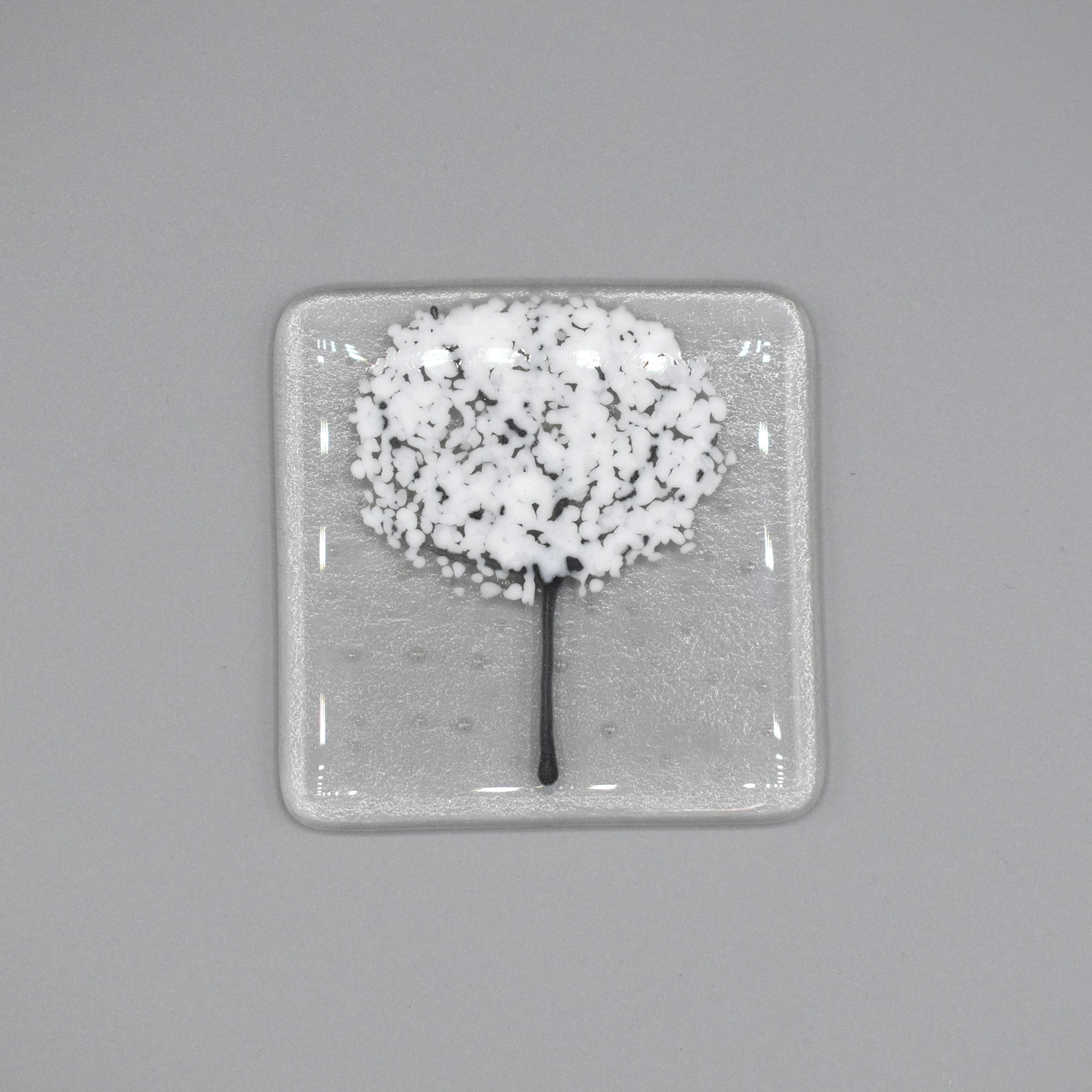 A clear glass square coaster with a white tree design