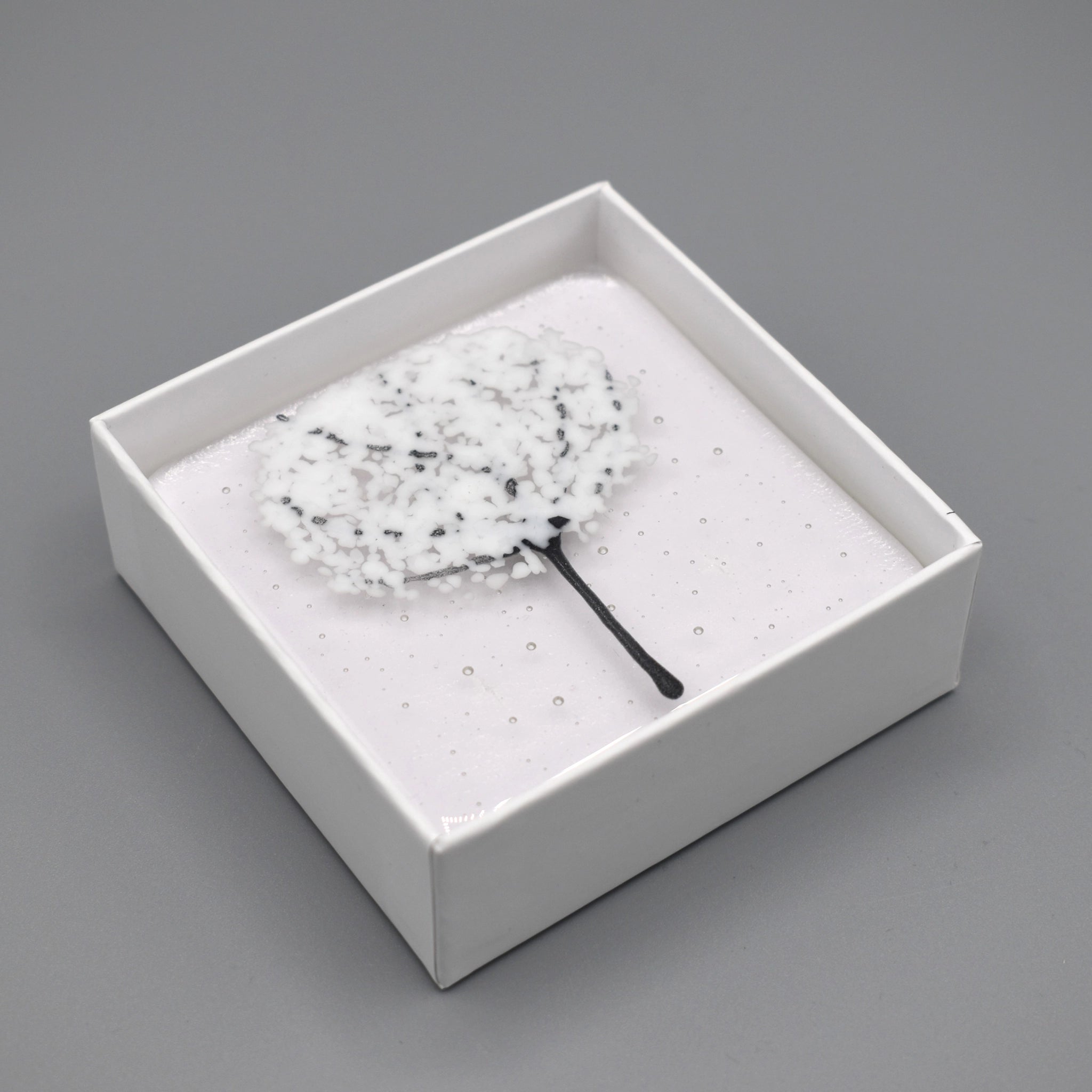 A clear glass square coaster with a white tree design in white box