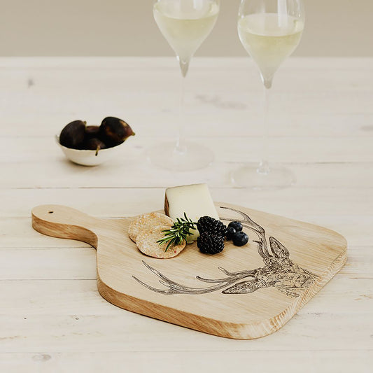 A wooden paddle style serving board with an engraved stag head staged on a table with cheese and crackers