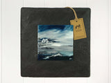 Wall art of a small white cottage next to a stormy sea in washed out blues and greys done in an abstract style onto a square ceramic tile mounted on square slate
