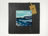Wall art of gentle waves in vibrant blues done in an abstract style onto a square ceramic tile mounted on square slate